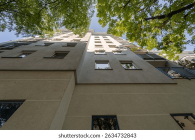 A low-angle perspective looking up the facade of a tall residential building. The view extends from the ground towards the sky, where the top of the building is framed by green tree leaves.