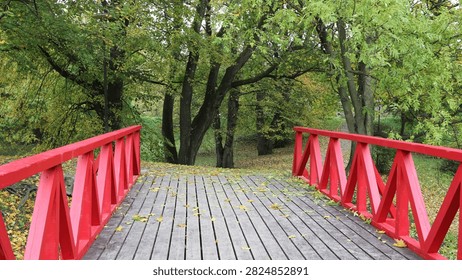 A low-angle perspective looking straight across a wooden footbridge featuring striking, bright red zig-zag railings. The weathered grey wooden planks of the walkway are lightly scattered with fallen yellow autumn leaves. The bridge leads directly into a tranquil park or forest area filled with lush green trees that are just beginning to show the early signs of fall foliage, creating a beautiful and vibrant contrast between the man-made structure and the natural environment.