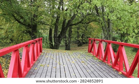 A low-angle perspective looking straight across a wooden footbridge featuring striking, bright red zig-zag railings. The weathered grey wooden planks of the walkway are lightly scattered with fallen yellow autumn leaves. The bridge leads directly into a tranquil park or forest area filled with lush green trees that are just beginning to show the early signs of fall foliage, creating a beautiful and vibrant contrast between the man-made structure and the natural environment.
