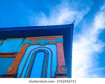 ​A low-angle perspective of a modern building facade in Bali featuring traditional orange brick pillars, tall arched windows, and white walls under a bright blue sky with wispy clouds.
