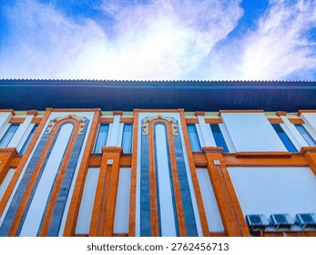 ​A low-angle perspective of a modern building facade in Bali featuring traditional orange brick pillars, tall arched windows, and white walls under a bright blue sky with wispy clouds.