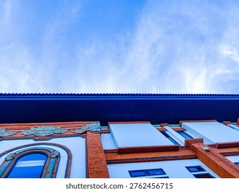 ​A low-angle perspective of a modern building facade in Bali featuring traditional orange brick pillars, tall arched windows, and white walls under a bright blue sky with wispy clouds.