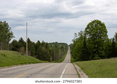 A low-angle perspective photograph of an empty, two-lane asphalt road dipping and rising into the distance through a quiet rural countryside or suburban woodland area. The worn pavement shows fine surface cracks, a yellow center line, and a faded white edge line, adding a rustic, realistic texture to the scene. Flanking the right side of the roadway is a prominent green tree canopy, while the left side features tall utility poles, power lines, and dense pine forests. The road leads the viewer's eye toward a straight incline on the distant horizon under a soft, overcast pale sky. This peaceful transportation scene perfectly captures the atmosphere of a classic American country highway, backroad route, or scenic drive. Ideal for high-quality commercial use in themes related to road trips, travel destinations, remote exploration, infrastructure development, and metaphors of journeys, pathways, or open roads.