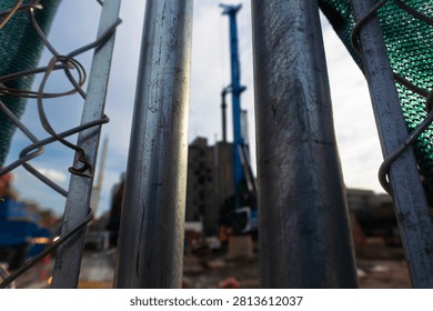 A low-angle, perspective shot looking through a weathered chain-link metal fence toward an active construction site. The blurred machinery and structures in the background convey the ongoing process of urban development and modernization in a bustling city environment, captured with an industrial, moody aesthetic.