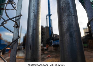 A low-angle, perspective shot looking through a weathered chain-link metal fence toward an active construction site. The blurred machinery and structures in the background convey the ongoing process of urban development and modernization in a bustling city environment, captured with an industrial, moody aesthetic.
