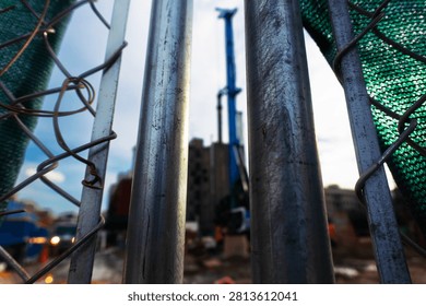 A low-angle, perspective shot looking through a weathered chain-link metal fence toward an active construction site. The blurred machinery and structures in the background convey the ongoing process of urban development and modernization in a bustling city environment, captured with an industrial, moody aesthetic.