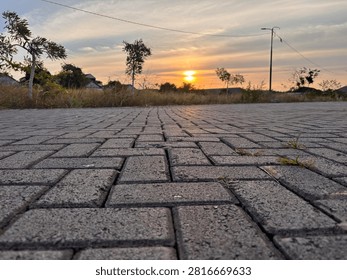 Low-angle photograph of an interlocking brick pavement leading toward a warm sunset over an open suburban landscape. The textured paving stones create strong leading lines and perspective, drawing the viewer’s eye toward the glowing horizon, scattered trees, and dramatic evening sky. Soft golden light enhances the peaceful atmosphere, while the combination of urban infrastructure and natural scenery makes this image suitable for themes of transportation, urban planning, outdoor lifestyle, travel, architecture, and nature. Ideal as a background with copy space for advertisements, presentations, websites, or editorial content.