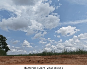 A low-angle, selective focus shot of a sandy rural path, with green grass and a solitary tree, contrasts the textured foreground with a vast blue sky filled with puffy clouds.