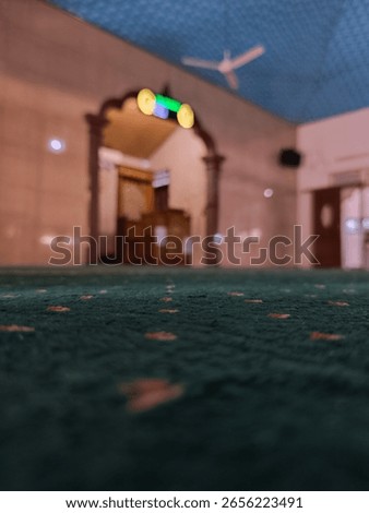 A low-angle, shallow depth-of-field shot of a mosque's green prayer carpet, with a blurred archway, mihrab, and ceiling fan in the background, conveying tranquility and spiritual ambiance.
