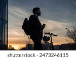 low-angle shot against backlight, a businessman enjoying a takeaway coffee with his electric scooter at dawn. The silhouette of the man and his electric vehicle stand out against the sunrise-lit sky
