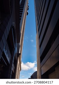 A low-angle shot between two tall buildings, highlighting their architectural lines against a bright blue sky with scattered clouds. The scene conveys a sense of urban density and perspective.