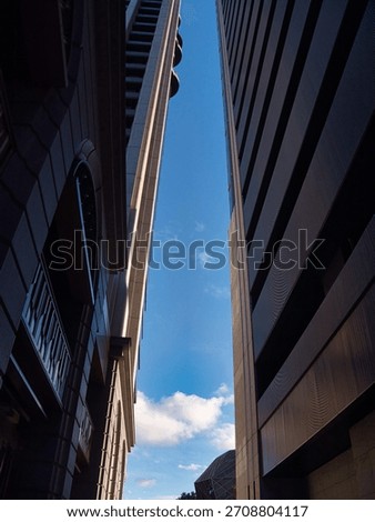 A low-angle shot between two tall buildings, highlighting their architectural lines against a bright blue sky with scattered clouds. The scene conveys a sense of urban density and perspective.