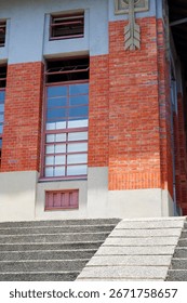 A low-angle shot of a building corner, featuring a red brick facade, tall windows, and a set of textured concrete stairs.