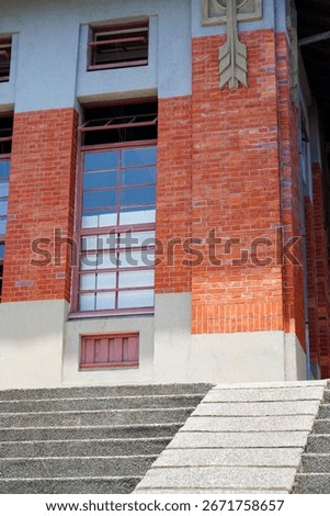 A low-angle shot of a building corner, featuring a red brick facade, tall windows, and a set of textured concrete stairs.
