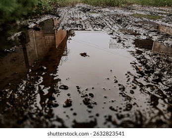 A low-angle shot captures a reflective puddle on a muddy track, mirroring distant buildings and fences, while tire tracks scar the foreground. Ideal for themes of weather, resilience, or urban decay.