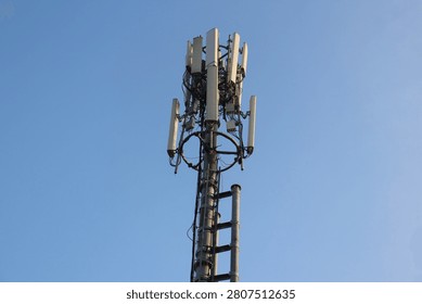 A low-angle shot of a cellular network tower or base transceiver station (BTS) equipped with multiple panel antennas and transmitters. The structure stands tall against a clear, bright blue sky background, representing mobile communication, wireless technology, 5G internet infrastructure, and global connectivity.