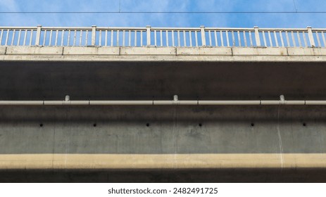 A low-angle shot of a concrete bridge underside, featuring a white railing, a pipe, and a clear blue sky above. Underneath the Bridge: A Concrete Perspective