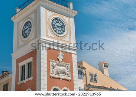 Low-angle shot of the historic clock tower featuring Roman numeral clock face and a Venetian Lion of St. Mark relief carving against a blue sky in Rovinj, Croatia.