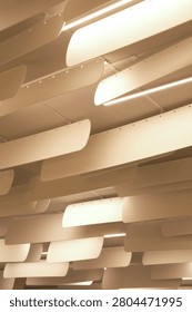 A low-angle shot looking up at a modern interior ceiling installation. The design features multiple rows of suspended, slightly curved, off-white panels hanging vertically in parallel lines, likely serving as acoustic baffles or architectural decorative elements. Integrated between these hanging panels are bright, linear LED light fixtures that cast a warm, soft glow across the space. The repetitive arrangement creates a striking, rhythmic geometric pattern and a strong sense of depth, characteristic of contemporary office, library, or commercial interior design