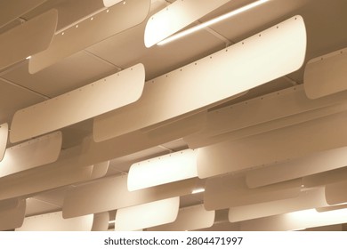 A low-angle shot looking up at a modern interior ceiling installation. The design features multiple rows of suspended, slightly curved, off-white panels hanging vertically in parallel lines, likely serving as acoustic baffles or architectural decorative elements. Integrated between these hanging panels are bright, linear LED light fixtures that cast a warm, soft glow across the space. The repetitive arrangement creates a striking, rhythmic geometric pattern and a strong sense of depth, characteristic of contemporary office, library, or commercial interior design