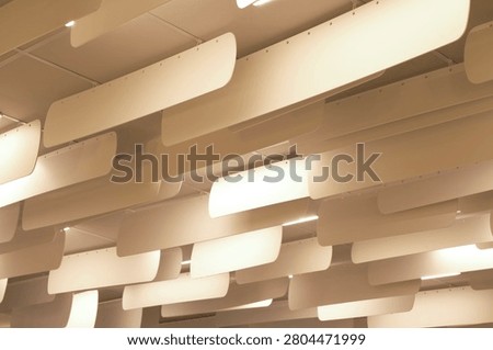 A low-angle shot looking up at a modern interior ceiling installation. The design features multiple rows of suspended, slightly curved, off-white panels hanging vertically in parallel lines, likely serving as acoustic baffles or architectural decorative elements. Integrated between these hanging panels are bright, linear LED light fixtures that cast a warm, soft glow across the space. The repetitive arrangement creates a striking, rhythmic geometric pattern and a strong sense of depth, characteristic of contemporary office, library, or commercial interior design