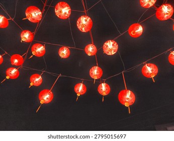 A low-angle shot looking up at numerous small, round red Chinese lanterns hanging from an outdoor wire network, illuminated brightly against the dark night sky.