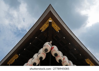 A low-angle shot looking up at the sharp, symmetrical eaves of a traditional Japanese building adorned with ornate gold details and a cluster of white paper lanterns against a cloudy sky.