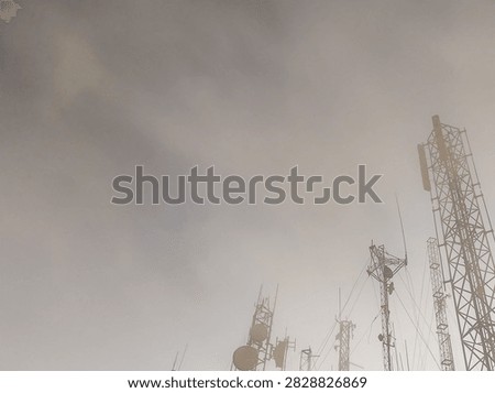 A low-angle shot of multiple cellular communication masts and radio towers shrouded in thick morning fog or smog. The structures appear silhouetted against a dense, hazy gray and overcast sky, symbolizing connectivity, infrastructure, and networking technology in bad weather.