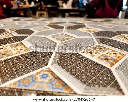 A low-angle shot of a table surface covered in an intricate Islamic geometric pattern featuring interlocking hexagons and octagons with diverse colors and repeating textile-like textures.