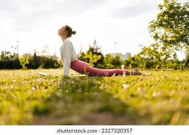 Low-angle side view of sportive and healthy young woman in white sportswear practicing yoga in green park doing Upward facing dog yoga pose on sporty mat. Concept of outdoors yoga, fitness and active. - Powered by Shutterstock