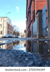 Low-angle street puddle reflection capturing parked cars, houses, autumn trees, and a bright sky with blue tones and white clouds, forming a mirrored urban scene.