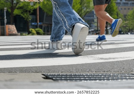 Low-angle of two people crossing intersection showcases motion. Captured mid-stride, highlights casual urban outfits - blue jeans and white platform sneakers and running wear and blue athletic shoes.