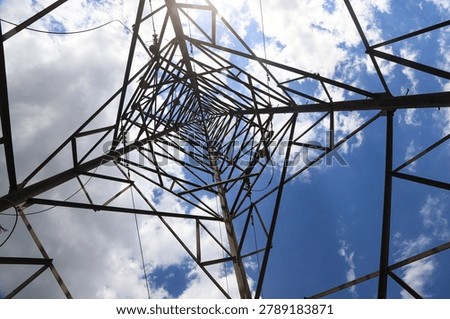 A low-angle upward shot showing the intersecting steel frame structure inside a high-voltage transmission tower, creating strong perspective lines and industrial geometric beauty against a blue sky with white clouds.