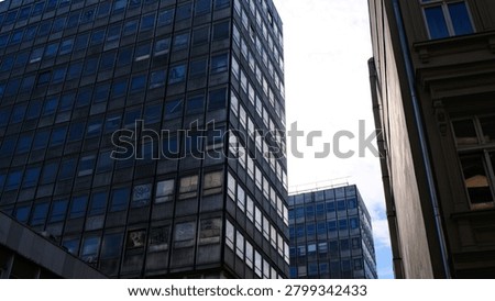 A low-angle, urban architectural shot featuring a cluster of aging, utilitarian office buildings, reminiscent of mid-century modern or brutalist design. The prominent high-rise structure dominates the frame with a rigid, repetitive grid of glass windows set into a dark, weathered facade. In the foreground, a lower, dilapidated building shows clear signs of urban decay, featuring peeling plaster and partially obscured or boarded-up windows. A second, similar high-rise stands in the background under a blue sky, highlighting a dense, stark cityscape