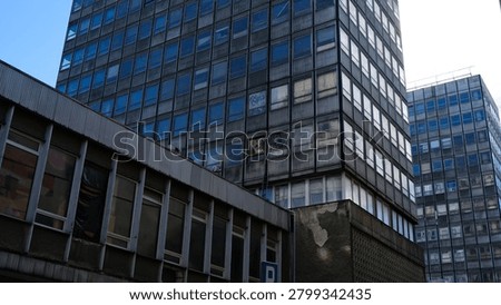 A low-angle, urban architectural shot featuring a cluster of aging, utilitarian office buildings, reminiscent of mid-century modern or brutalist design. The prominent high-rise structure dominates the frame with a rigid, repetitive grid of glass windows set into a dark, weathered facade. In the foreground, a lower, dilapidated building shows clear signs of urban decay, featuring peeling plaster and partially obscured or boarded-up windows. A second, similar high-rise stands in the background under a blue sky, highlighting a dense, stark cityscape