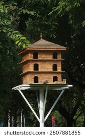 A low-angle vertical shot of a beautiful multi-tier wooden birdhouse or dovecote built like a traditional pagoda. Elevated on a white metal pole surrounded by lush green trees in a public park, perfect for wildlife, garden decoration, and nature concepts.