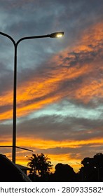 A low-angle, vertical shot captures a vibrant sunset over a silhouetted suburban landscape. On the left side of the frame, a prominent dark grey streetlamp curves over, its light bulb glowing brightly with a warm white illumination. The sky behind it is filled with dynamic, textured clouds painted in brilliant shades of fiery orange and golden-yellow from the setting sun, contrasting with layers of heavy, dark blue-grey clouds. Near the horizon, the silhouettes of tropical palm trees and dense foliage stand out. In the lower left foreground, a small section of a white vehicle's roof and side window is visible, anchoring the viewpoint.