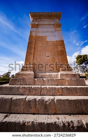 A low-angle, vertical shot capturing the full height and imposing structure of the Roman funerary tower. The massive stepped base leads up to the smooth-faced tower body, all set against a vibrant blue sky.