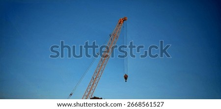 Low-angle view of a bright orange construction crane set against a clear blue sky. The crane’s lattice structure and suspended hook highlight its engineering detail and industrial strength. 