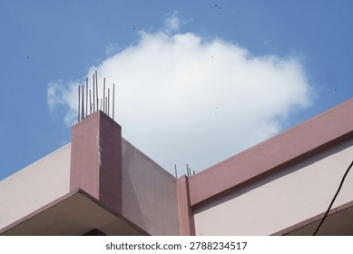 A low-angle view of a building corner under construction, featuring exposed rebar against a bright blue sky with a fluffy white cloud, indicating ongoing development.