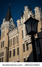A low-angle view captures the intricate architecture of the National Bank of Ukraine building in Kyiv, featuring its distinctive cream and white facade, multiple turrets, and a prominent spire topped with a weather vane. A dark streetlamp stands in silhouette in the foreground, complementing the historical urban scene under a bright, clear blue sky.