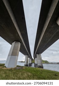A low-angle view captures a massive concrete bridge spanning across a body of water, supported by geometric pillars under an overcast sky.