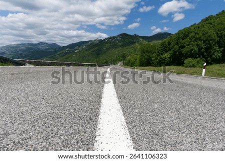 A low-angle view captures a paved road stretching towards distant green mountains under a partly cloudy sky, with a white center line guiding the eye.
