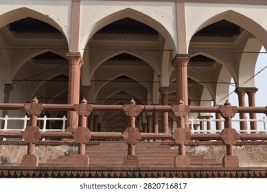 A low-angle view captures a red sandstone carved balustrade railing in the immediate foreground, leading up to an open-air pavilion structure. Multiple rows of pointed arches and supporting columns stretch into the background under soft, natural daylight.
