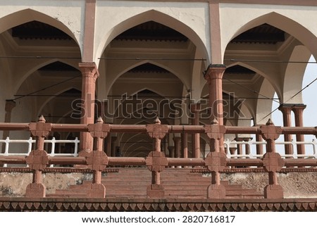 A low-angle view captures a red sandstone carved balustrade railing in the immediate foreground, leading up to an open-air pavilion structure. Multiple rows of pointed arches and supporting columns stretch into the background under soft, natural daylight.