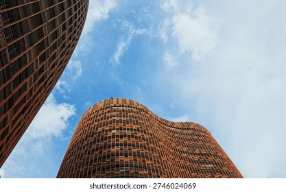 A low-angle view captures two distinct modern skyscrapers with striking wavy, curved facades and a grid pattern in warm orange, yellow, and red tones, set against a clear blue sky dotted with white clouds. 