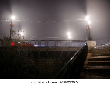 A low-angle view capturing a red car driving across a bridge on a foggy night, its taillights creating streaks. Streetlights illuminate the mist and the metal railing, 