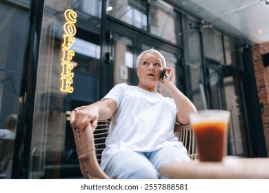 Low-angle view of casual senior woman with surprised expression sitting in outdoor cafe, holding mobile phone to ear, while takeaway coffee sitting on table in front of her. - Powered by Shutterstock