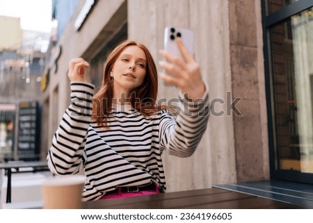 Low-angle view of charming redhead female tourist making selfie or video call to friends sharing impressions of summer trip. Attractive young woman holding smartphone in hand, video calling.