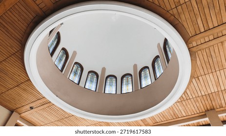 Low-angle view of a circular architectural dome with stained glass windows set into a wooden plank ceiling. Modern interior design with elegant lighting and symmetry. High quality.